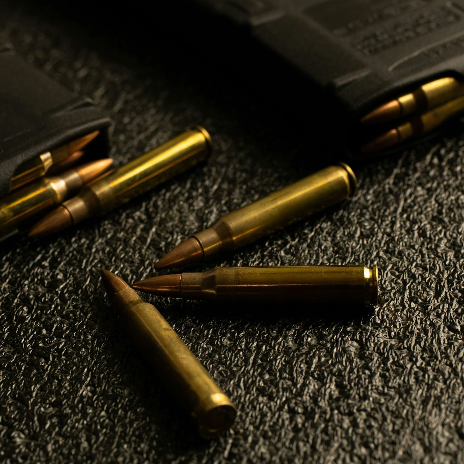 A close-up of rifle cartridges lying on a textured black surface, with some rounds partially loaded in black magazines.
